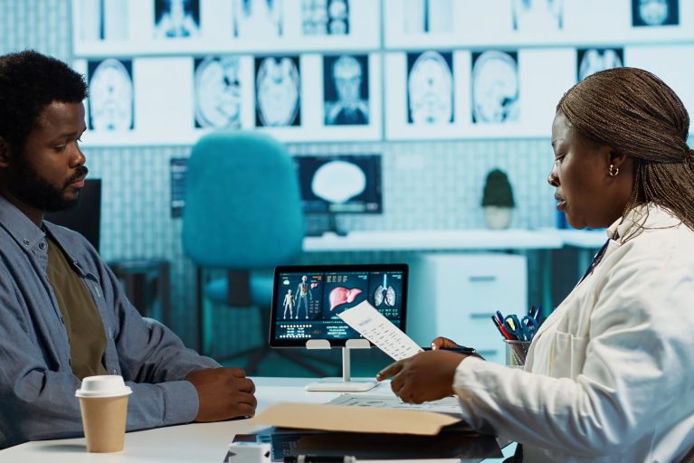 Female physician using radiology scans and reports to advise patient during checkup focusing on diagnosis and treatment. African american doctor with medical expertise consults casual man. Camera A.